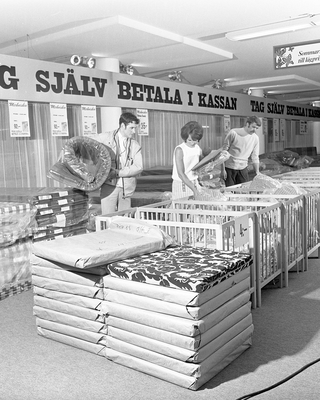 Customers selecting and packing items in an IKEA store with a sign about paying at the checkout