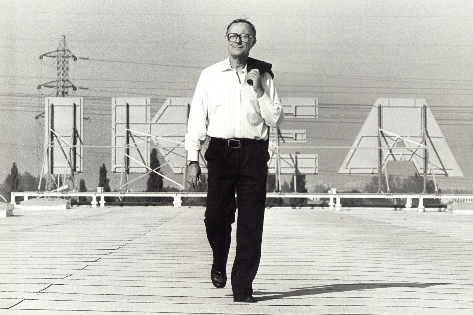 Ingvar Kamprad walking on a rooftop with an IKEA sign in the background, wearing a shirt and glasses, with a relaxed posture