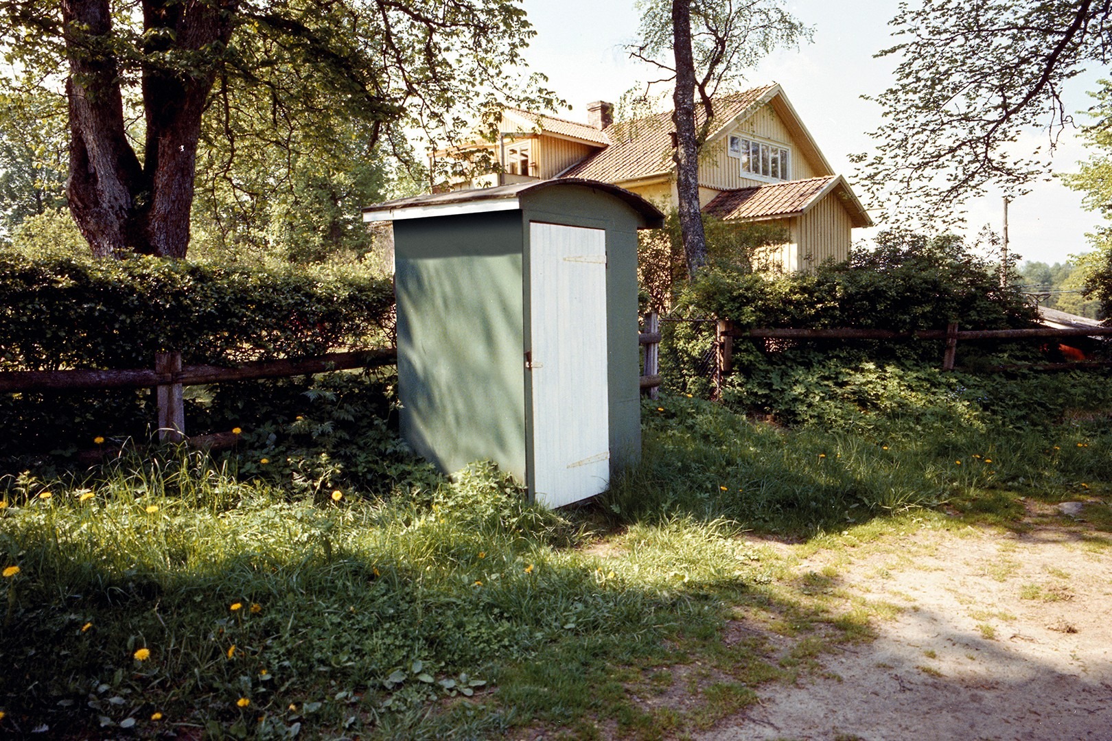 Small green shed in a garden with grass and trees, with a yellow wooden house in the background