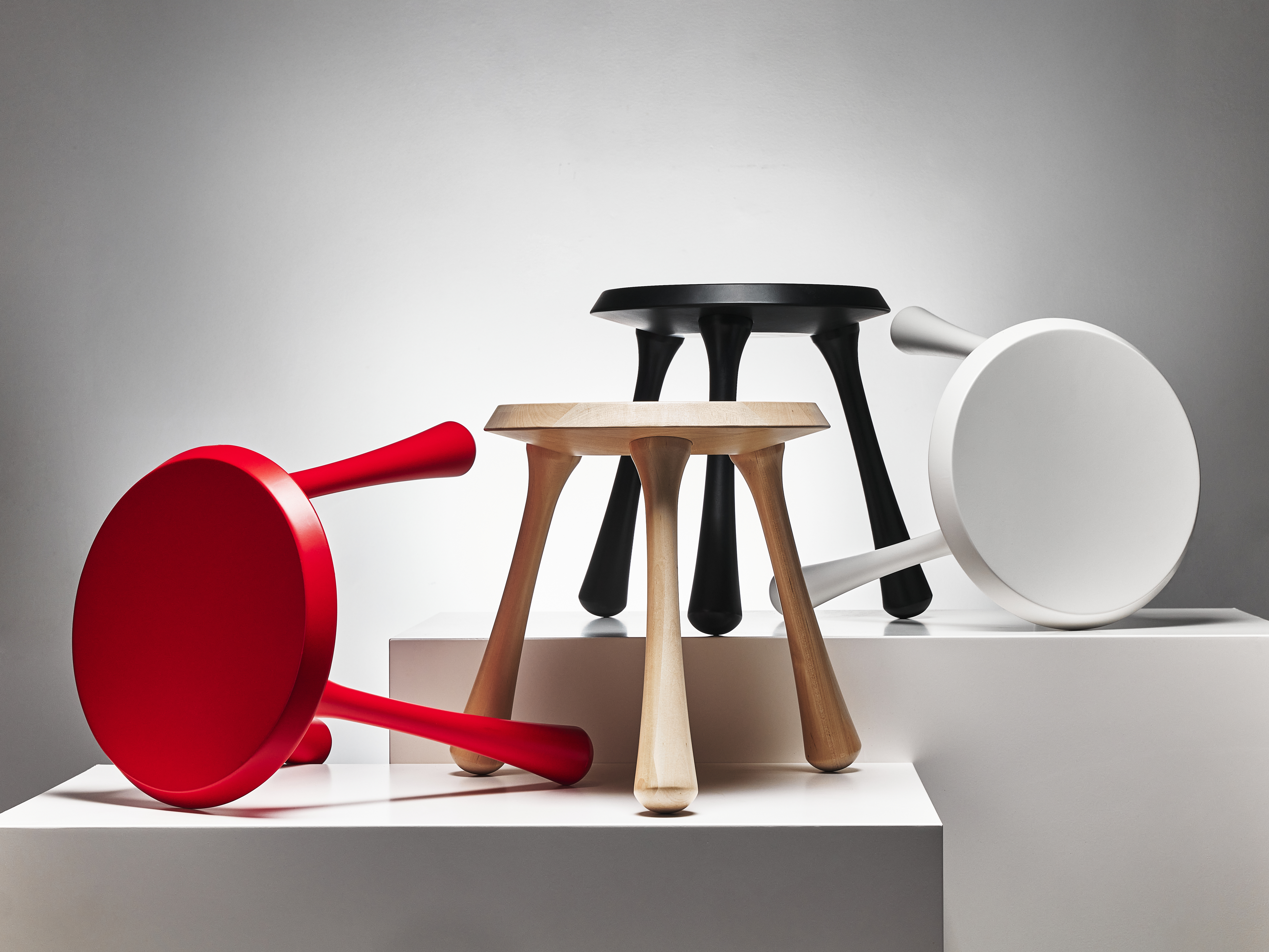 Four stools in different colors displayed on white pedestals.